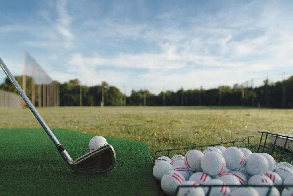 a driving range with a small basket of golf balls on the right side of picture, green and a club getting ready to hit a golf ball along with trees in the background and blue skies